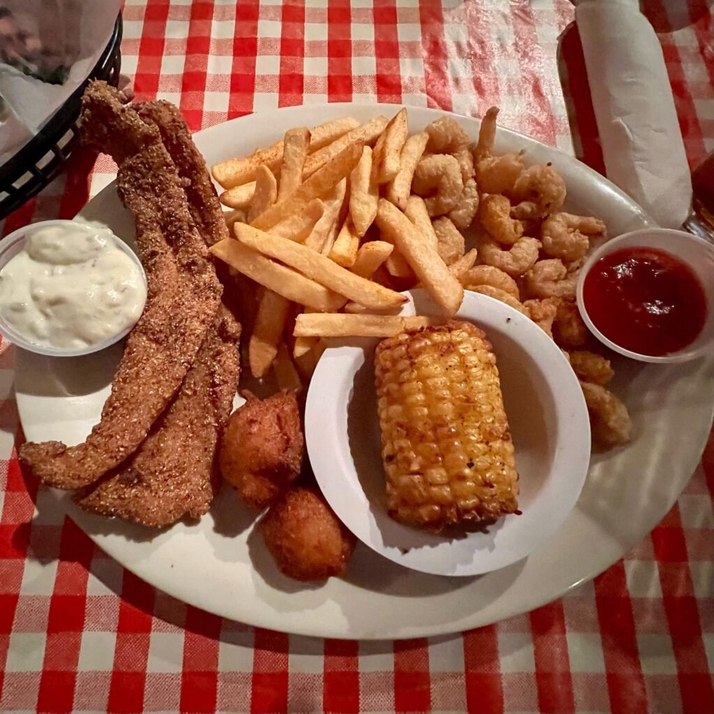 A platter loaded with fried fish, french fries, hush puppies, corn, and dipping sauces served on a red and white checkered tablecloth.