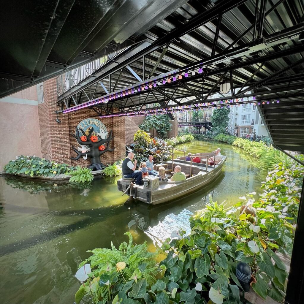 Passengers enjoy a boat trip under an iron bridge surrounded by lush greenery. The scene captures the old-world charm and architecture of the covered canal.