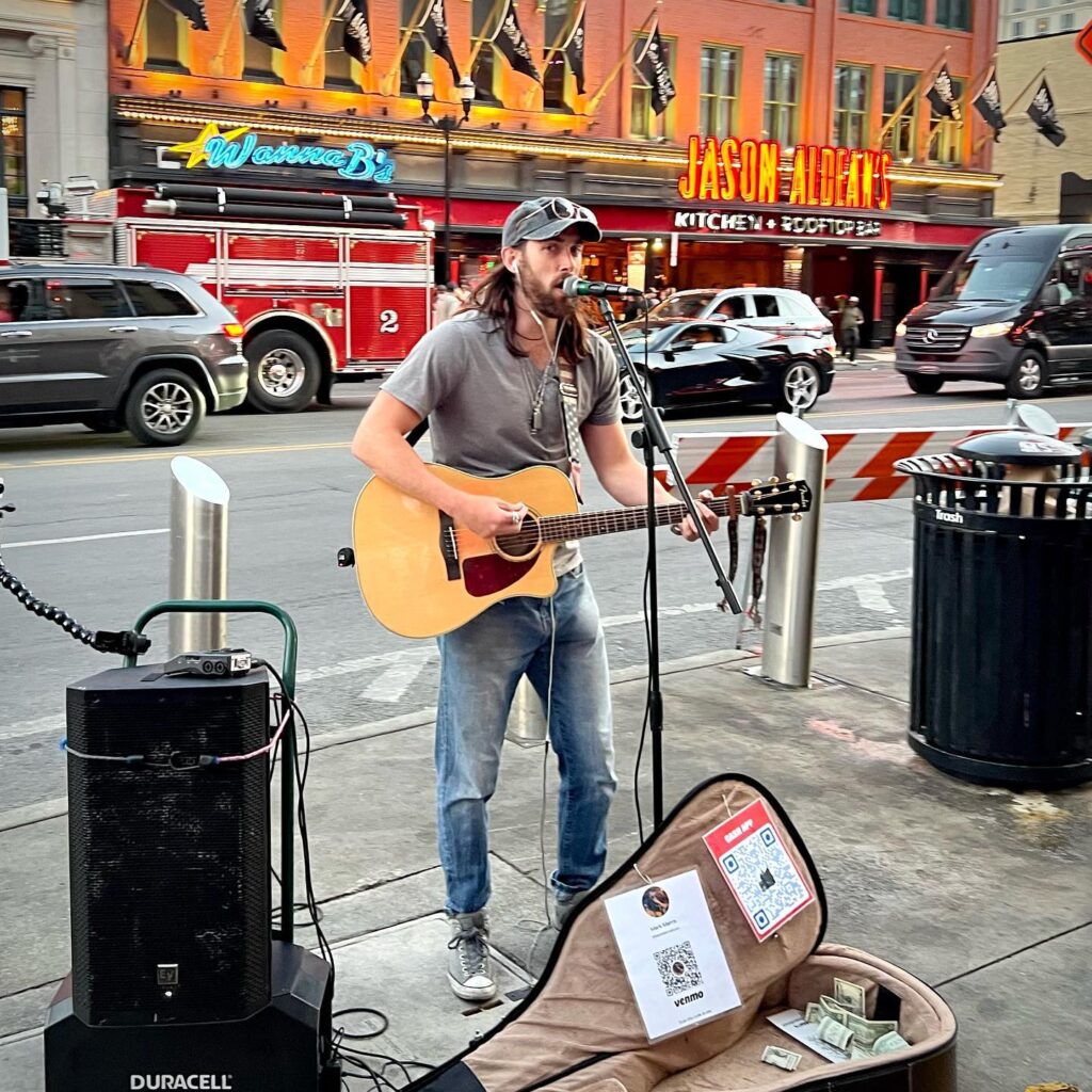 A musician performs busking on a city sidewalk in front of the Jason Albert Restaurant on Pike Place in Seattle. A guitar case with a QR code is open on the ground for tips.