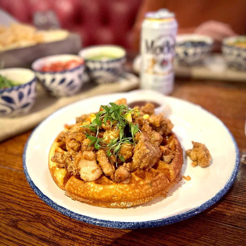 A plate of savory fried chicken and waffles garnished with fresh microgreens. Small condiment bowls and a beverage can are visible in the background.