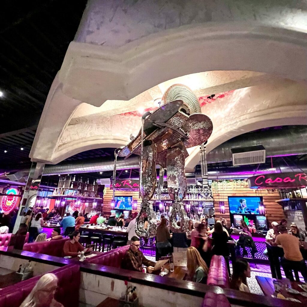 A retro-styled diner interior with a large artistic installation on the ceiling. Pink neon lights and busy tables create a lively scene.