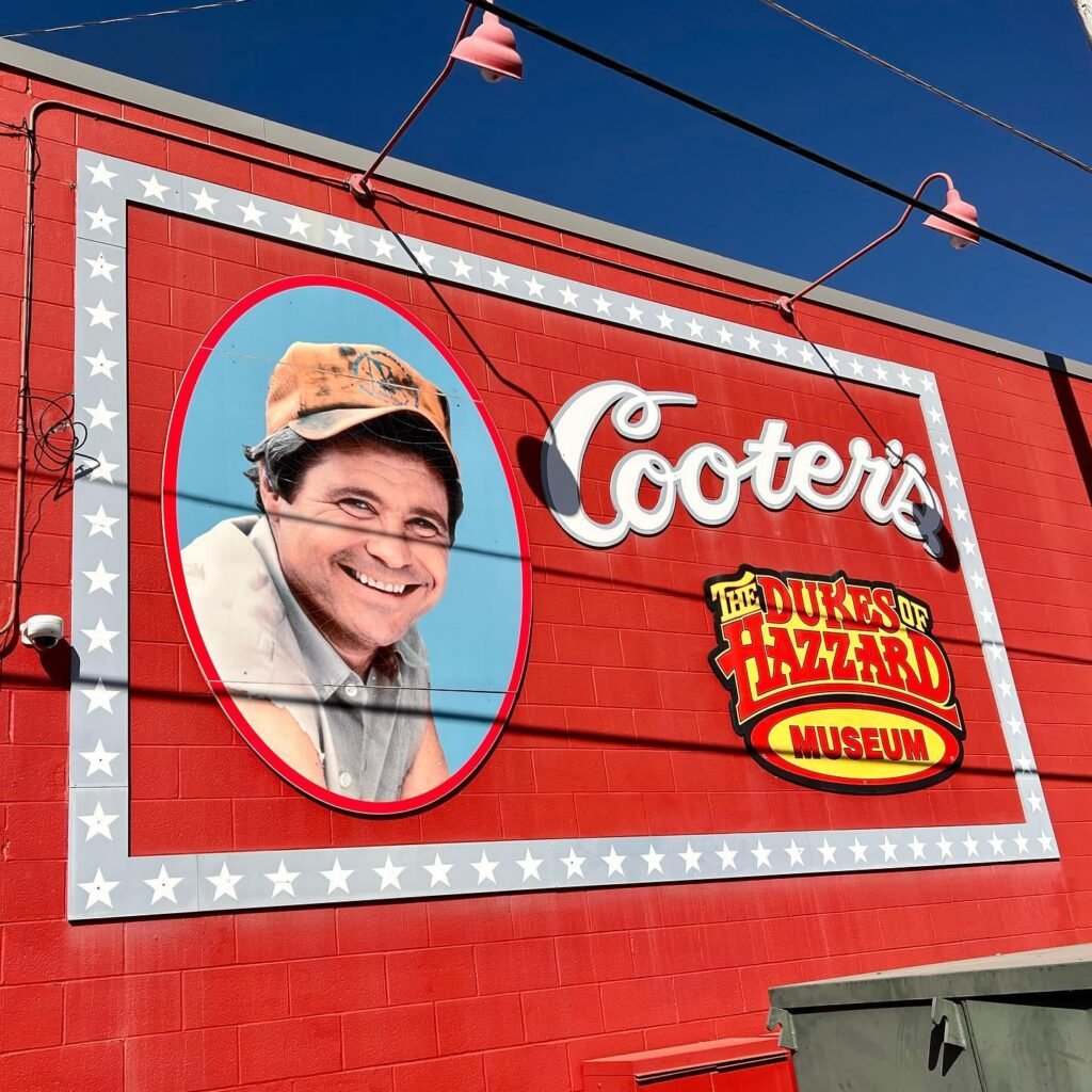 A red brick building features a large mural of Cooter's and the Dukes of Hazzard Museum logo. A smiling man in a cap is prominently displayed within the artwork.