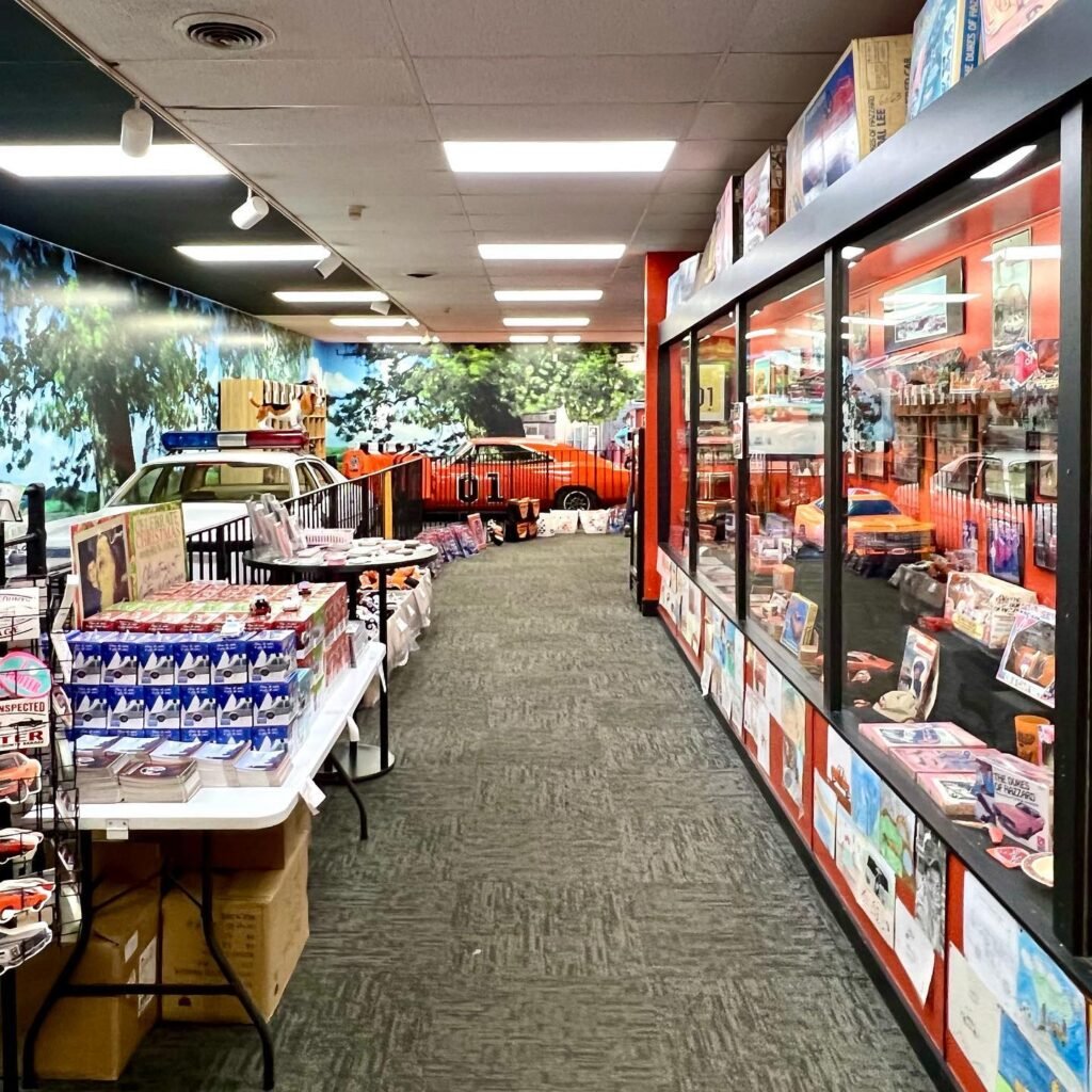 View of a long aisle in a store with tables and glass display cases lined with magazines and postcards, featuring an automotive-themed mural on the far wall.
