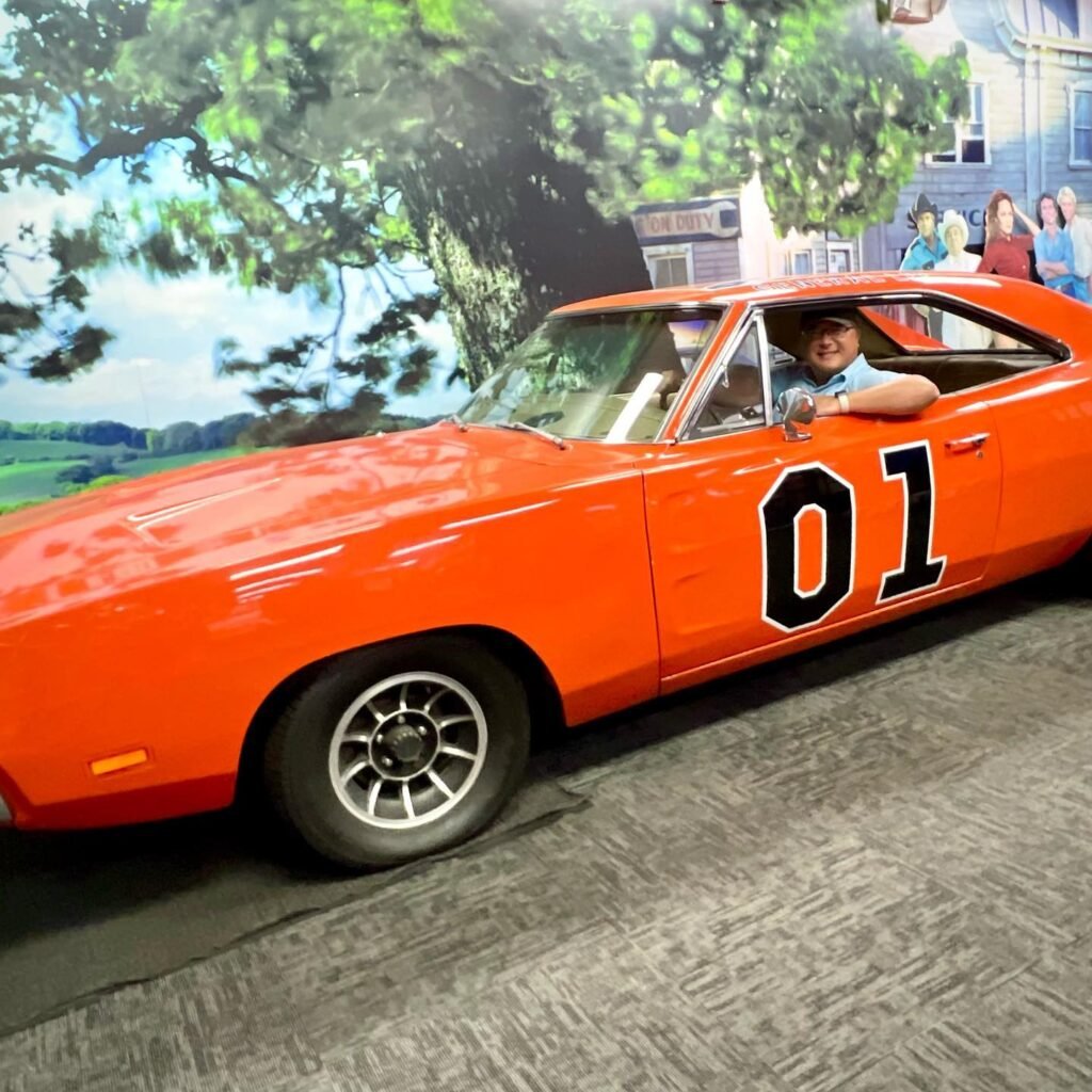 An orange 1978 Pontiac Firebird Trans Am with the number 01 on the door. A person is driving a replica vehicle in front of a movie mural.