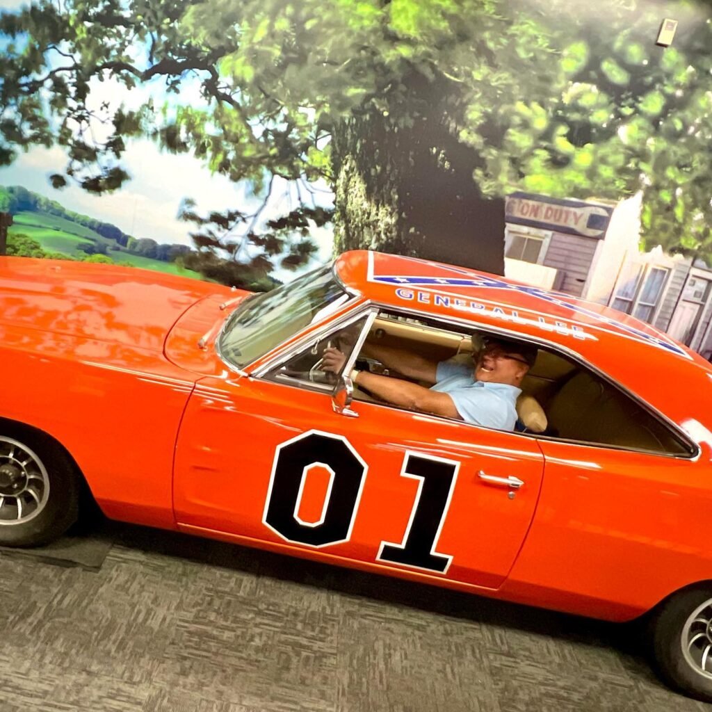 A bright orange muscle car marked with the number 01 is displayed on a themed trade show booth. The backdrop features scenic landscapes and buildings to set the scene.
