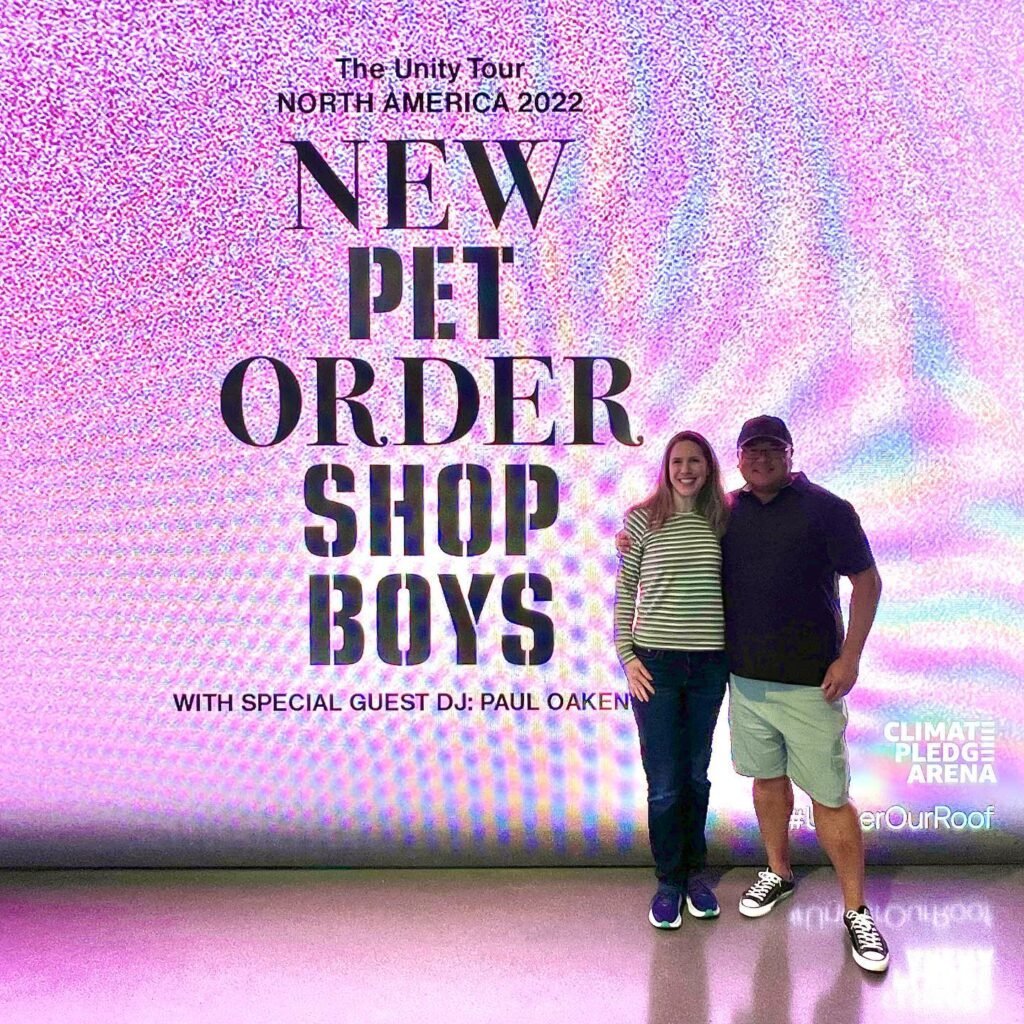 Two people posing in front of a promotional backdrop for The New Order Shop Boys North America 2022 tour at Climate Pledge Arena.