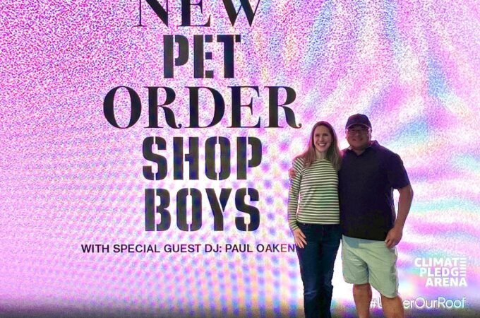 Two people posing in front of a promotional backdrop for The New Order Shop Boys North America 2022 tour at Climate Pledge Arena.