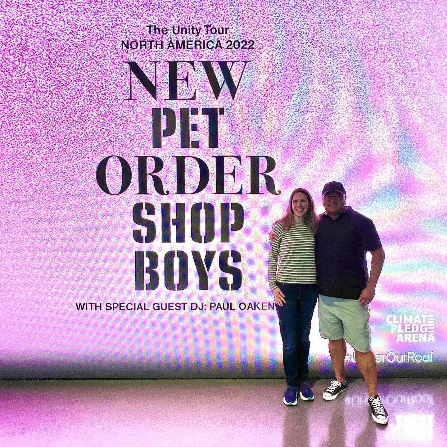 Two people posing in front of a promotional backdrop for The New Order Shop Boys North America 2022 tour at Climate Pledge Arena.