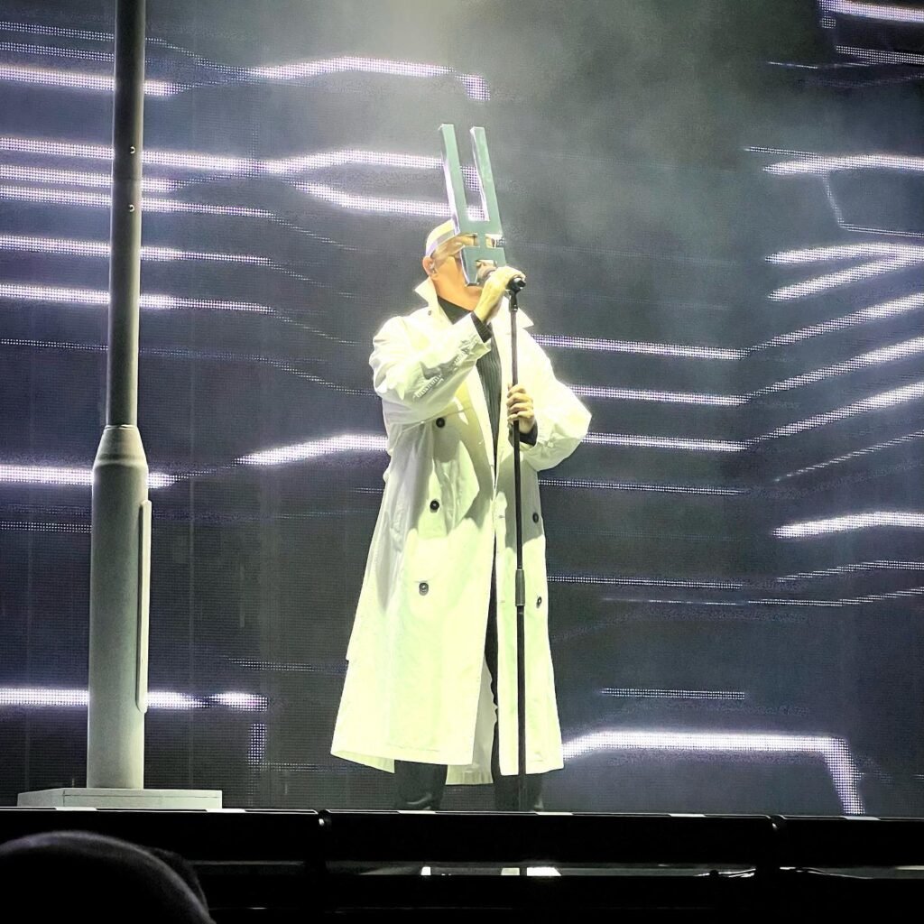 A performer wearing a white coat stands on stage holding a microphone stand.