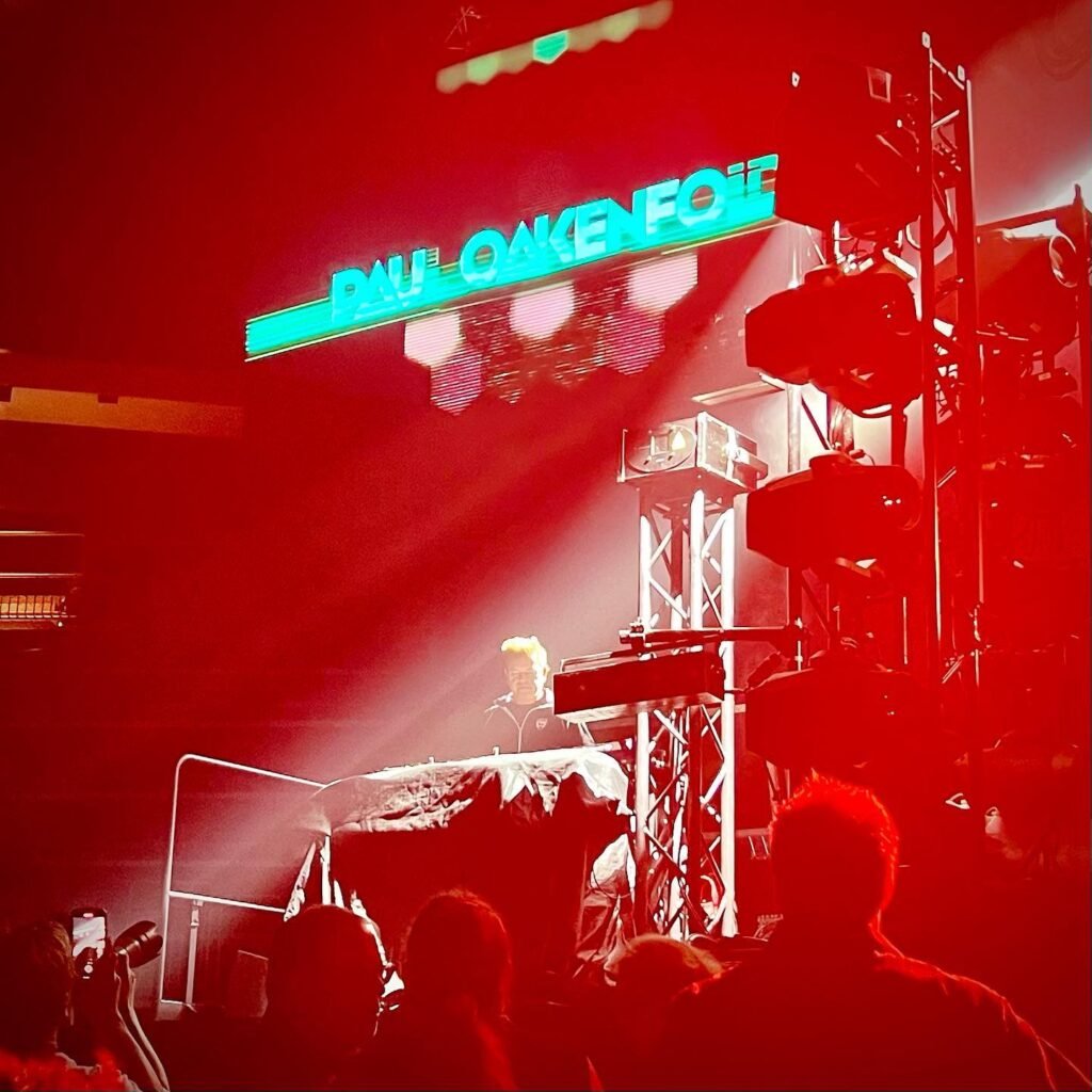 A DJ performing on stage under red lights with a Paul Oakenfold neon sign.