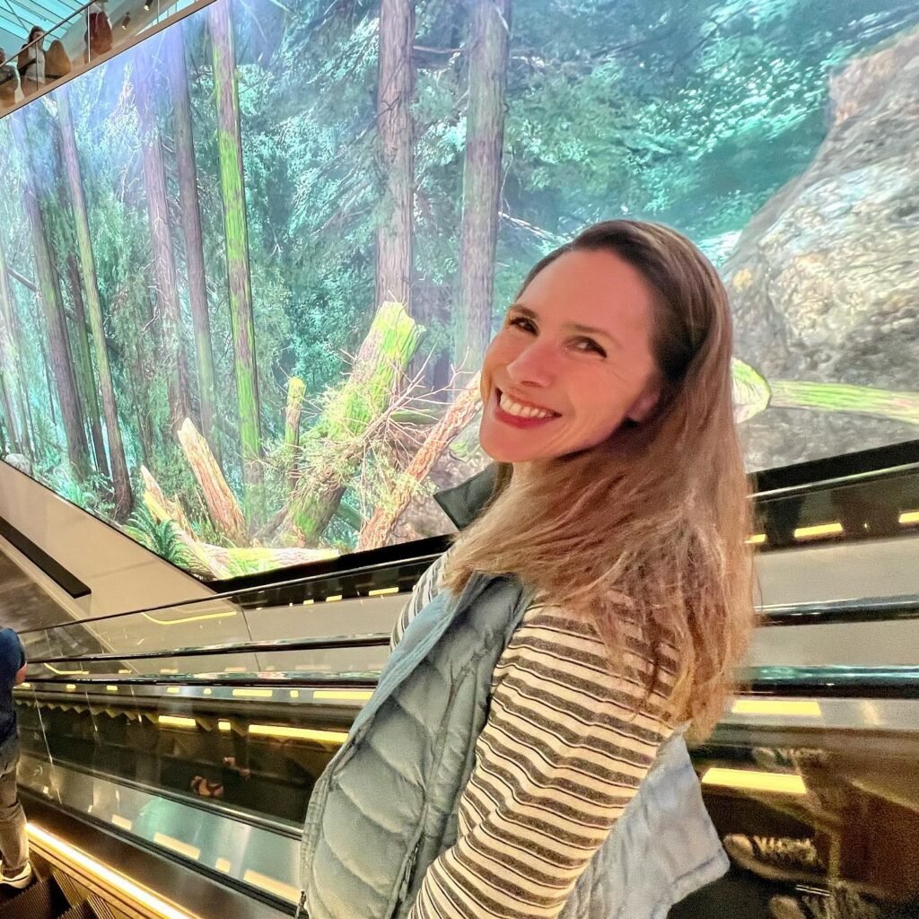 A smiling visitor takes a photo in front of a large mural depicting a Pacific Northwest rainforest. The person wears a striped shirt and a puffy vest at the amusement park.