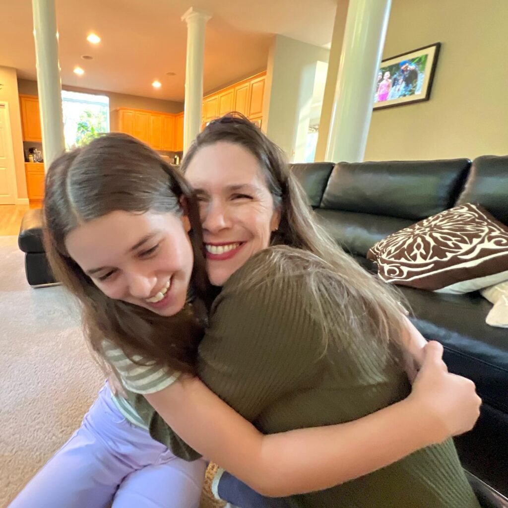 A joyful hug between two smiling individuals in a warmly lit living room. The background includes a black leather sofa, a decorative pillow, and a framed family photo on the wall.
