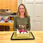 A happy woman in a green sweater sits behind a sheet cake with four lit candles in a home kitchen.
