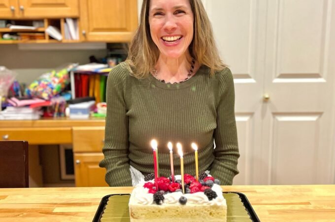 A happy woman in a green sweater sits behind a sheet cake with four lit candles in a home kitchen.