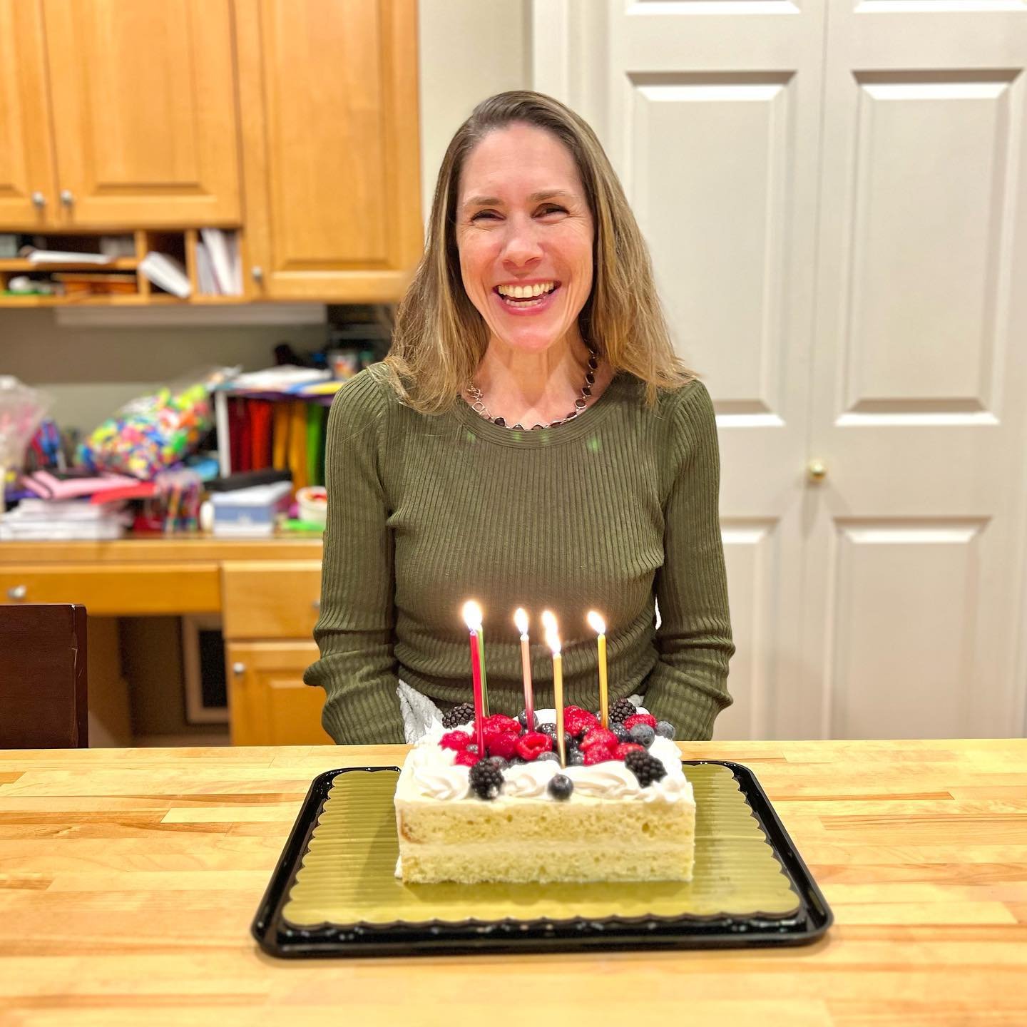 A happy woman in a green sweater sits behind a sheet cake with four lit candles in a home kitchen.