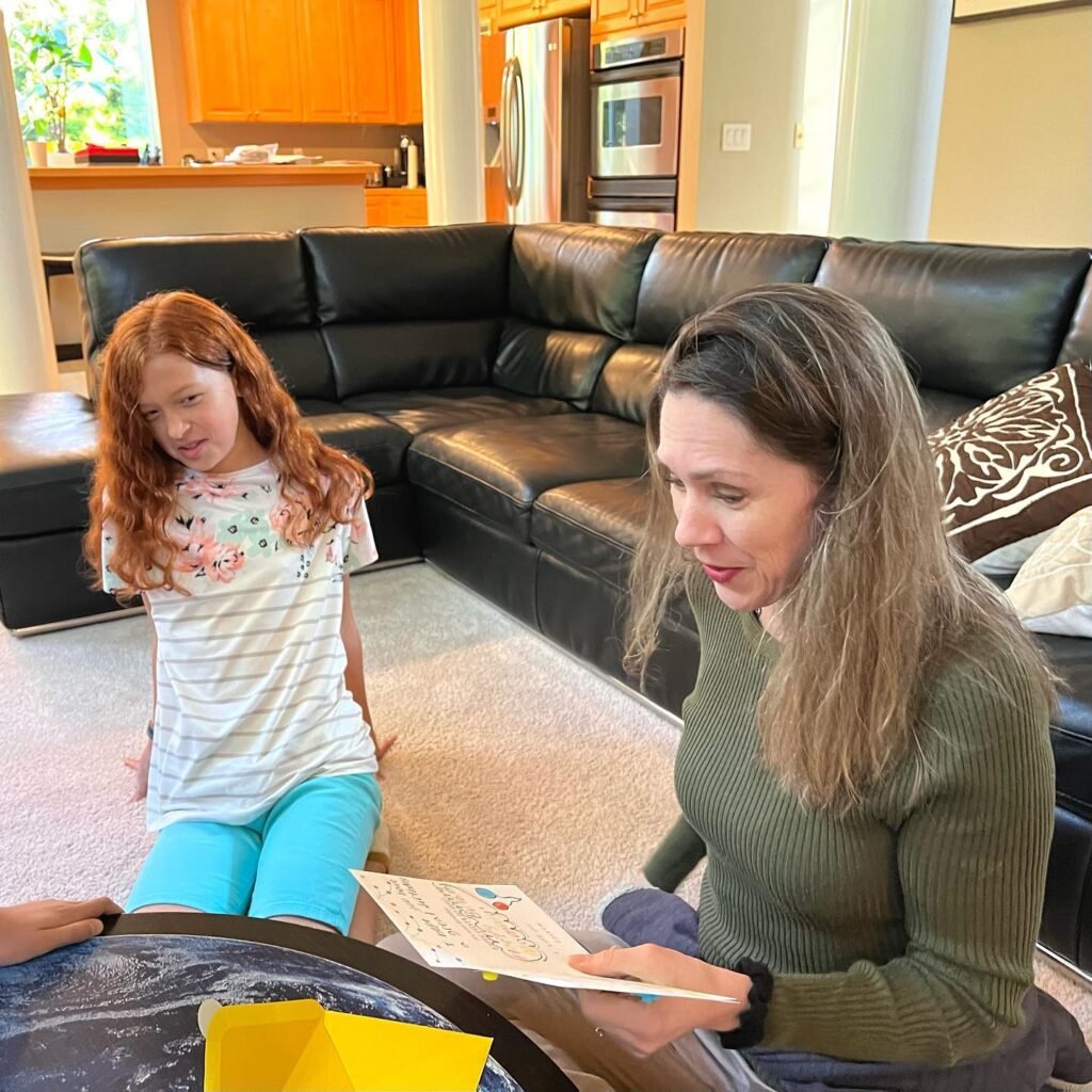 A collage showing two people engaged in reading and discussing a book in a home setting. The background features a modern living area with a black leather sofa and wooden cabinets.