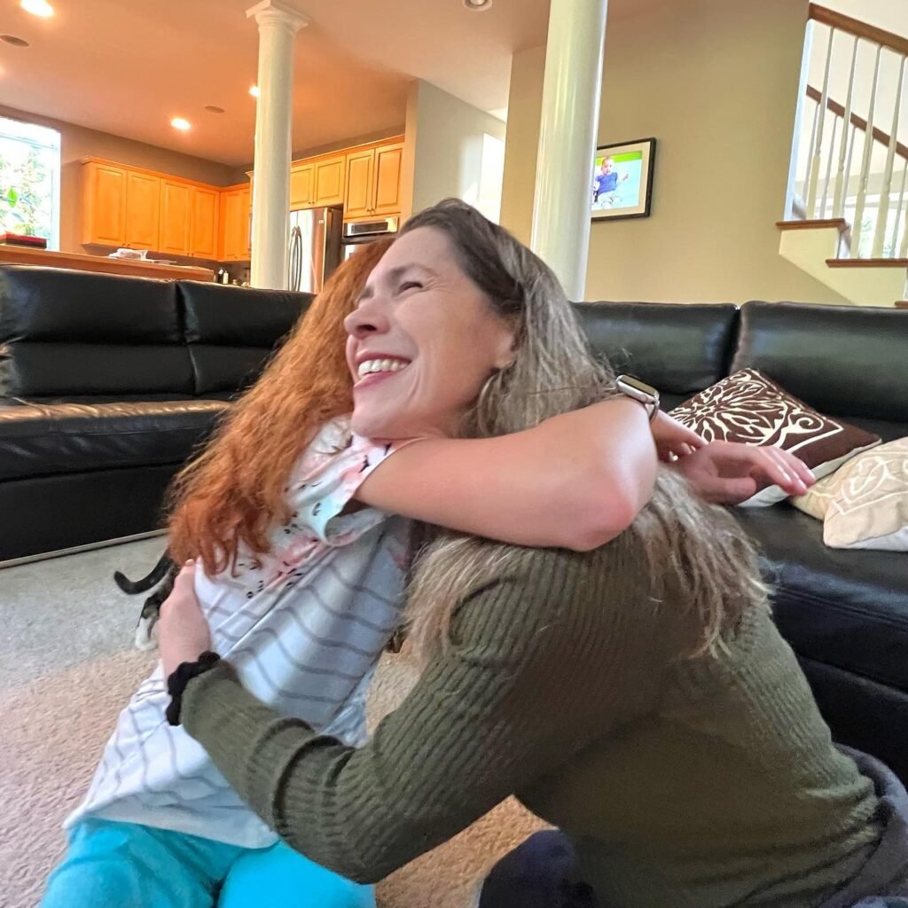 A warm embrace between a red-haired child and an adult in a homey living room featuring wooden kitchen cabinets and a black leather sofa.