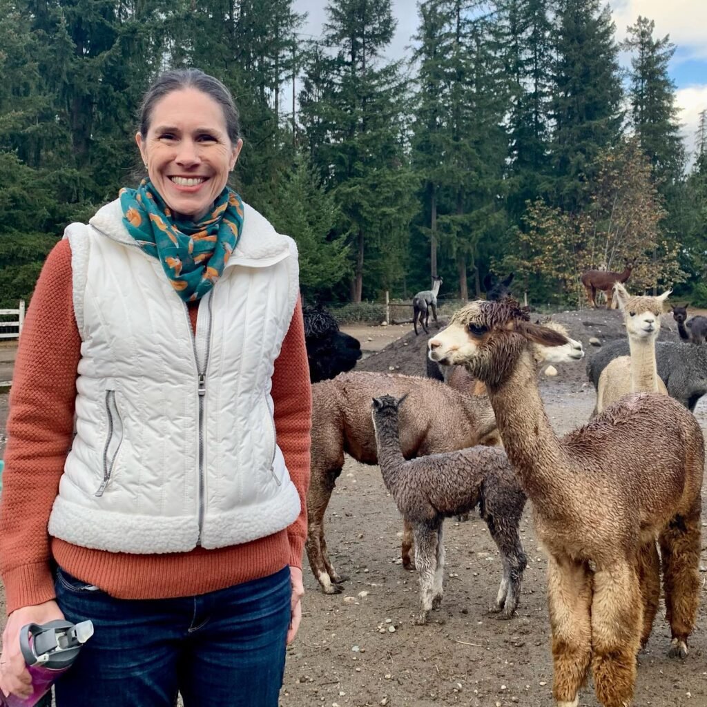 A smiling smile woman in a white vest and teal scarf standing near alpacas in a forest clearing.