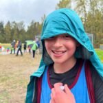A smiling student wearing a teal hooded raincoat in front of an outdoor event field with a forest backdrop.