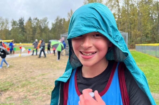 A smiling student wearing a teal hooded raincoat in front of an outdoor event field with a forest backdrop.