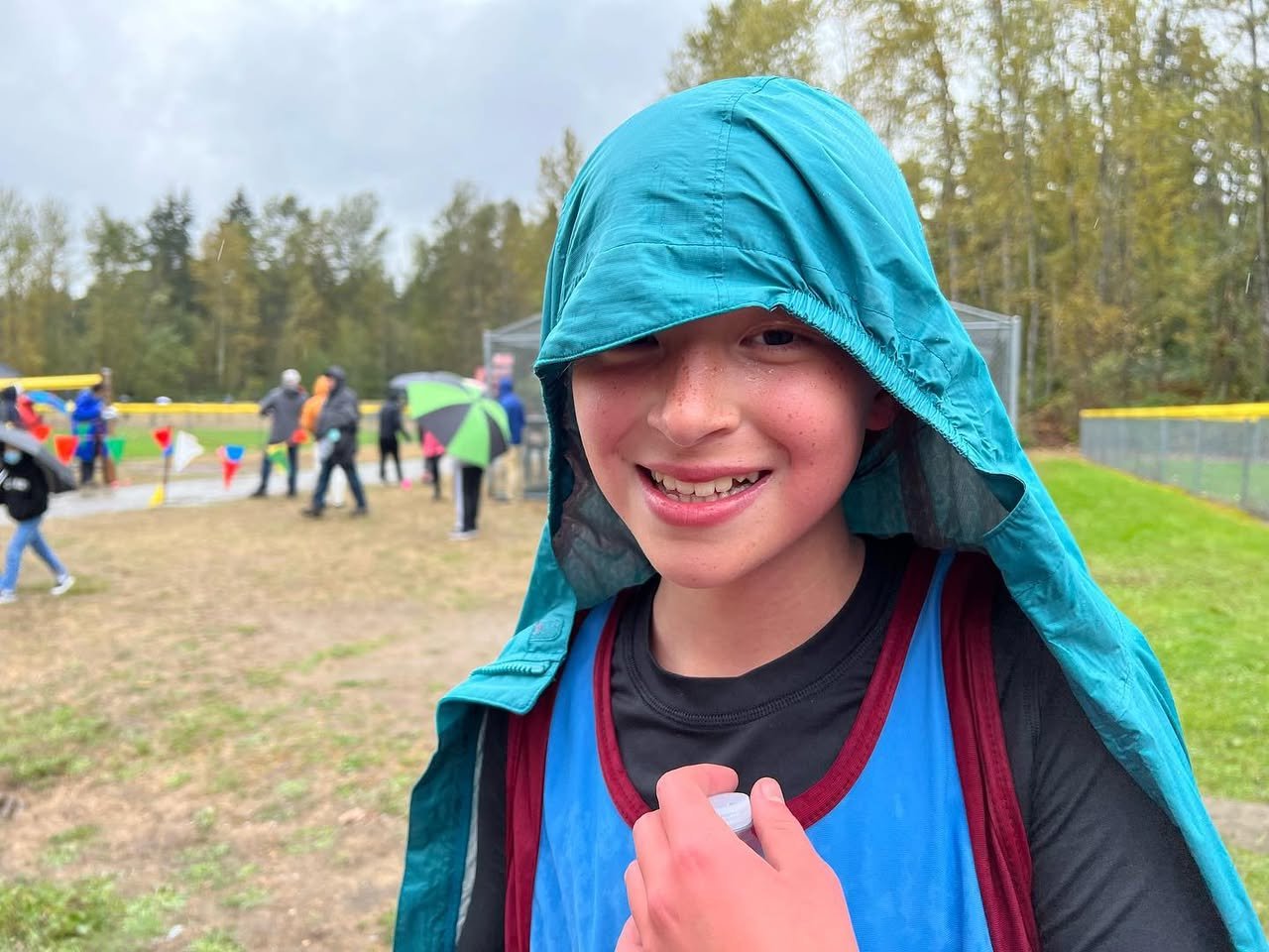 Big Finn Hill Park A smiling student wearing a teal hooded raincoat in front of an outdoor event field with a forest backdrop.