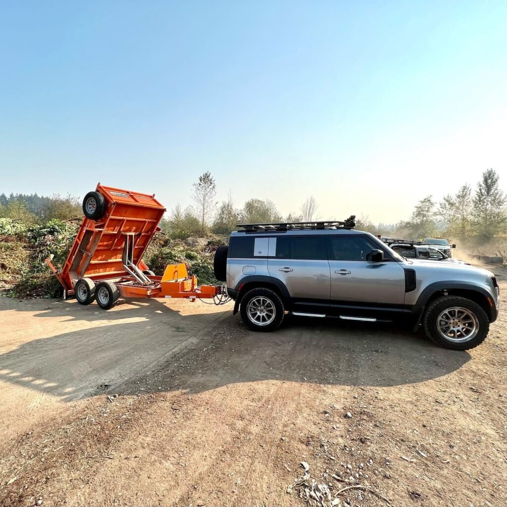 A silver SUV is towing an orange dump trailer in a rural setting. The trailer's bed is tilted, ready for unloading materials on a dirt path.