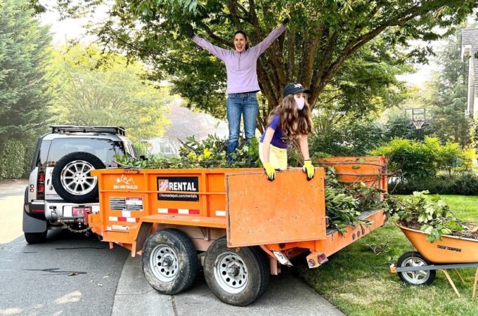 Two people stand on top of a large orange trailer filled with green tree branches.