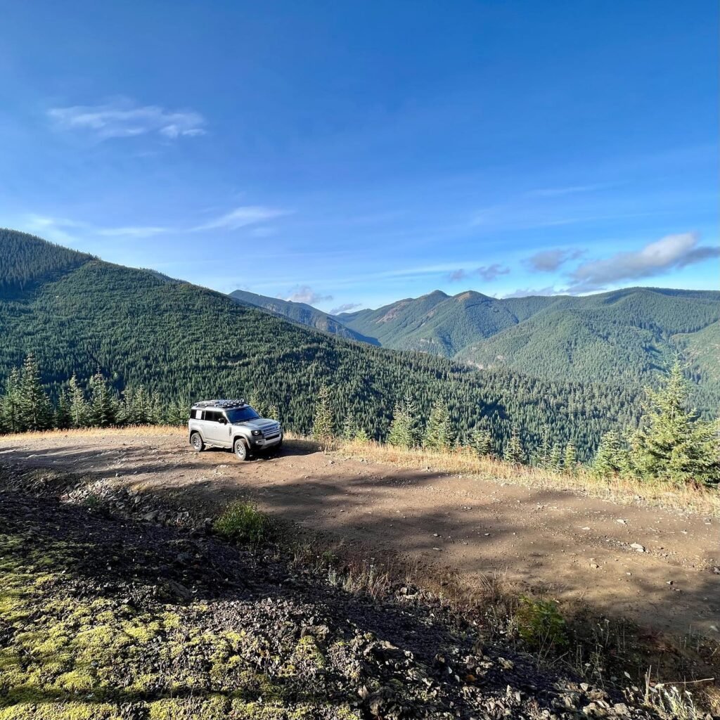 A scenic view of a white vehicle parked on a dirt overlook at a mountain peak, surrounded by dense evergreen forest.