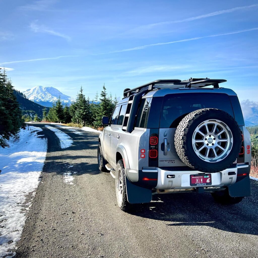 A silver SUV parked on a mountain road beside patches of snow, with a snow-capped peak in the background. The scene shows a sunny day in the Pacific Northwest with evergreen trees.