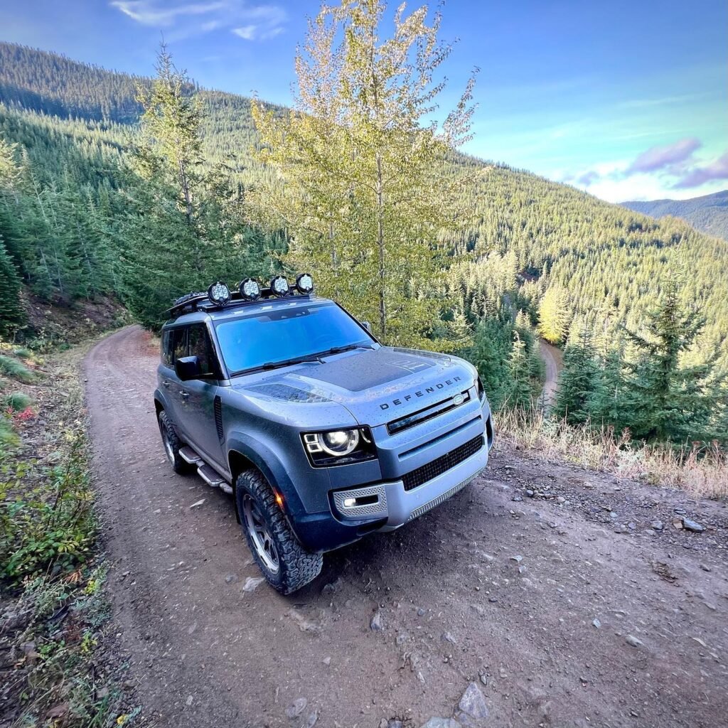 A grey SUV with a roof-light bar navigating a dirt trail through a lush mountain forest in the Pacific Northwest.