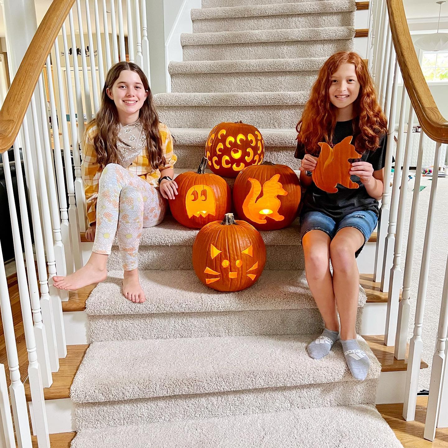 Two girls sit on a carpeted staircase posing with lit Jack-o-Lanterns during a Halloween family gathering.