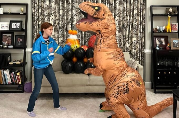 A child in a blue jacket playing with a large, inflatable T-Rex dinosaur costume in a living room.