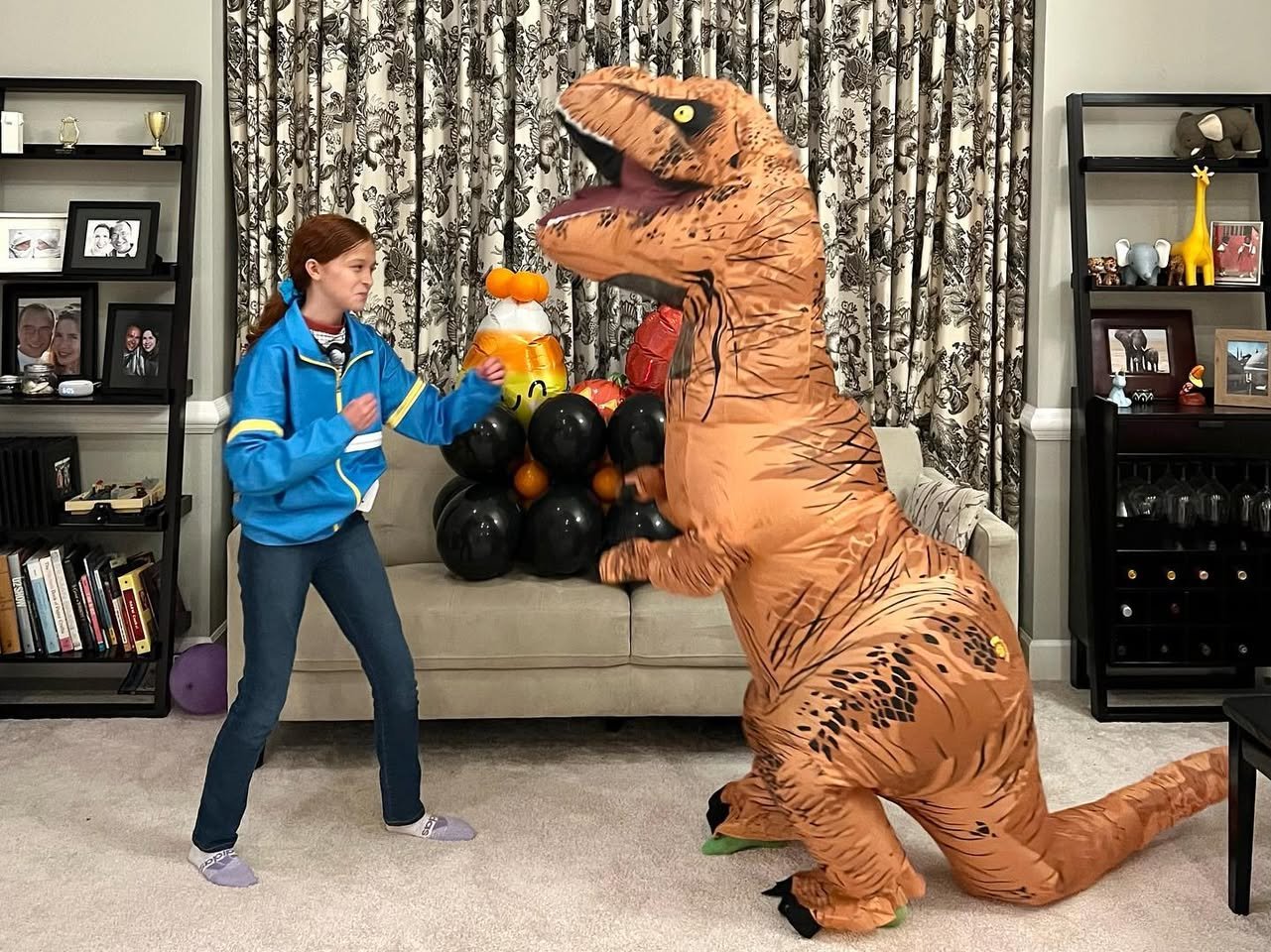 A child in a blue jacket playing with a large, inflatable T-Rex dinosaur costume in a living room.