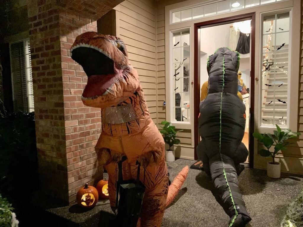 An indoor and outdoor view of inflatable T-Rex and Apatosaurus figures on a porch at night. The dinosaur is next to a glowing jack-o-lantern pumpkin.