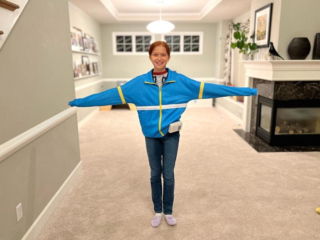 A person standing in a home hallway with arms outstretched wearing a blue Cocomotion vest. Below is a close-up view of the blue vest sleeves and the indoor carpet.