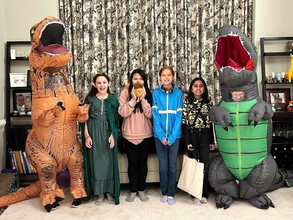 Four people posing with two large inflatable dinosaur costumes in a living room featuring a patterned curtain background and bookshelves.