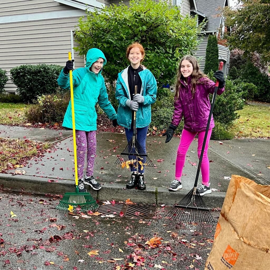 Three young people wearing colorful raincoats and rain pants use leaf rakes to clear a driveway scattered with orange and red autumn leaves.