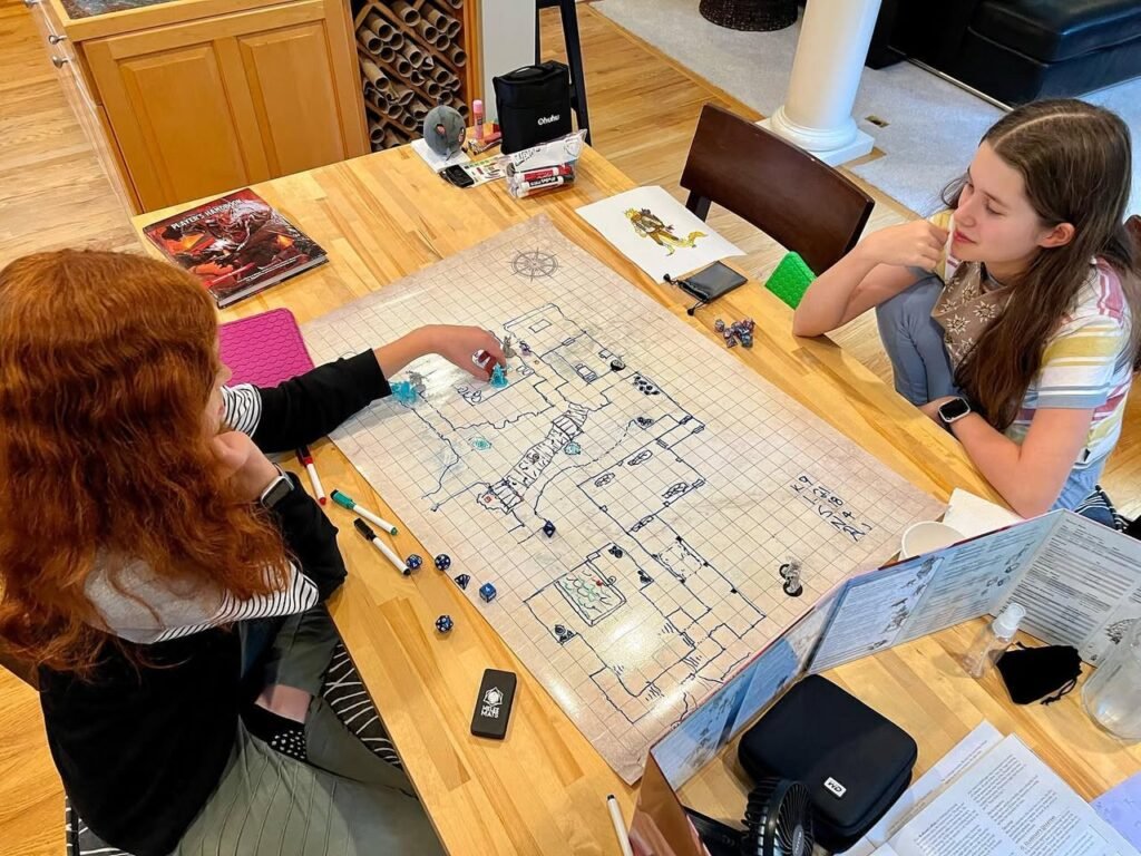 Two people play a board game with a detailed map layout on a wooden table. The game involves placing various tokens and markers on the grid.