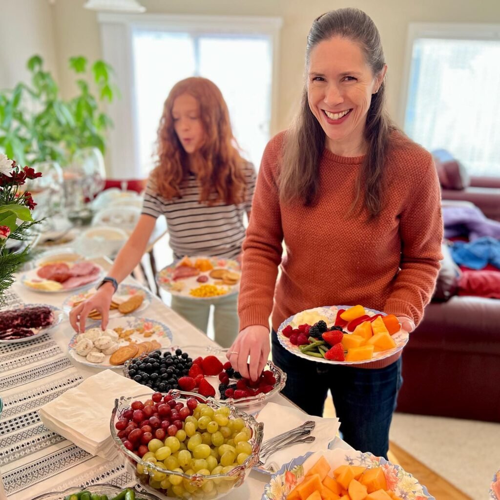 Two people are standing at a dining table filled with bowls of fresh fruit and charcuterie. A smiling woman in an orange sweater holds a plate of colorful berries.