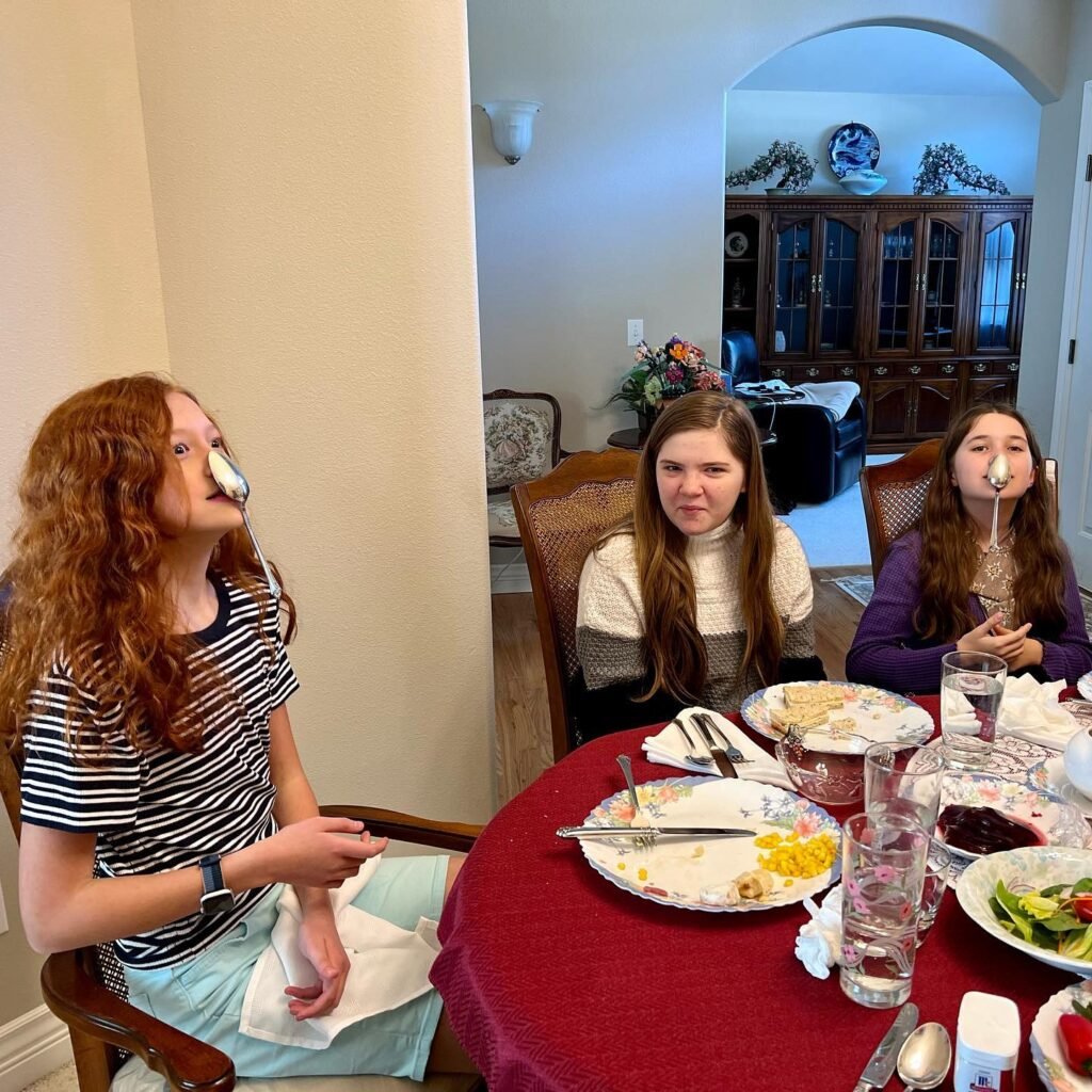 A family enjoys a silly dinner together at a table set with a red tablecloth. Dinnerware is visible on the table in the warm dining room.