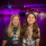Two girls smiling in front of a roller rink with purple lighting.