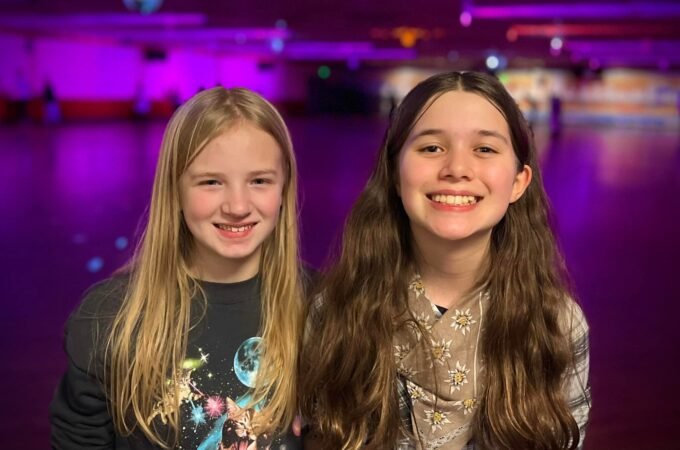 Two girls smiling in front of a roller rink with purple lighting.