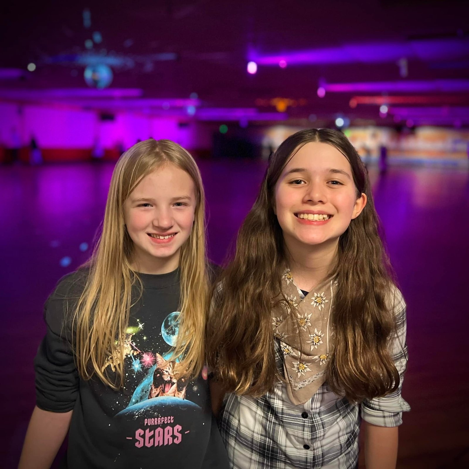 Two girls smiling in front of a roller rink with purple lighting.