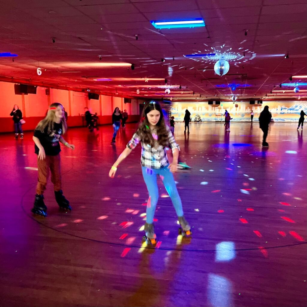 People enjoying roller skating in a brightly lit indoor rink with colorful reflections on the floor. The scene captures the lively atmosphere of a family-friendly recreational activity.