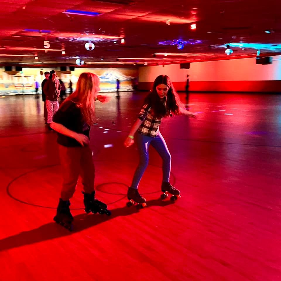 People roller skating in a vibrant indoor rink illuminated by bright red lighting. The scene is lively and energetic with colorful reflections on the smooth surface.