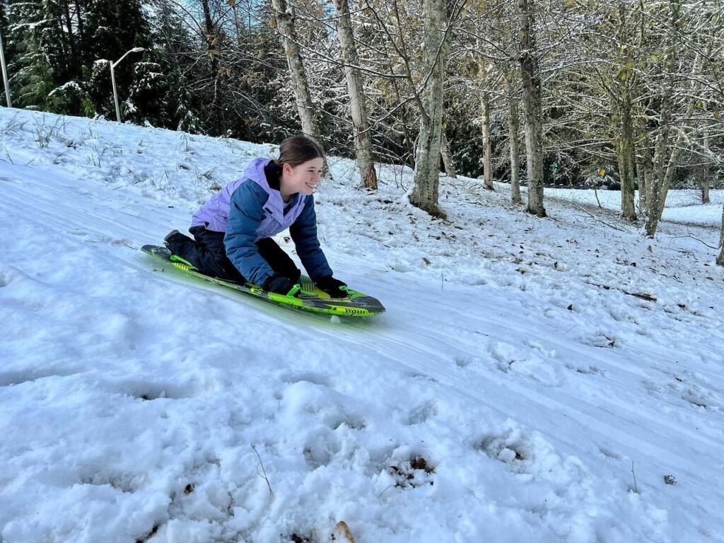 A person slides down a snowy hill on a green plastic sled, surrounded by leafless trees in the Pacific Northwest woods. The trail is packed with snow, capturing a winter moment.