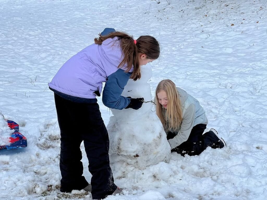 Two people constructing a snowman in a snowy field. One person kneels shaping the base while the other stands nearby.