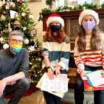 A small group wearing Santa hats and face masks practice singing near a beautifully decorated Christmas tree, holding sheet music. They are in a warm, festive indoor setting.