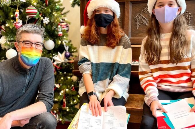 A small group wearing Santa hats and face masks practice singing near a beautifully decorated Christmas tree, holding sheet music. They are in a warm, festive indoor setting.