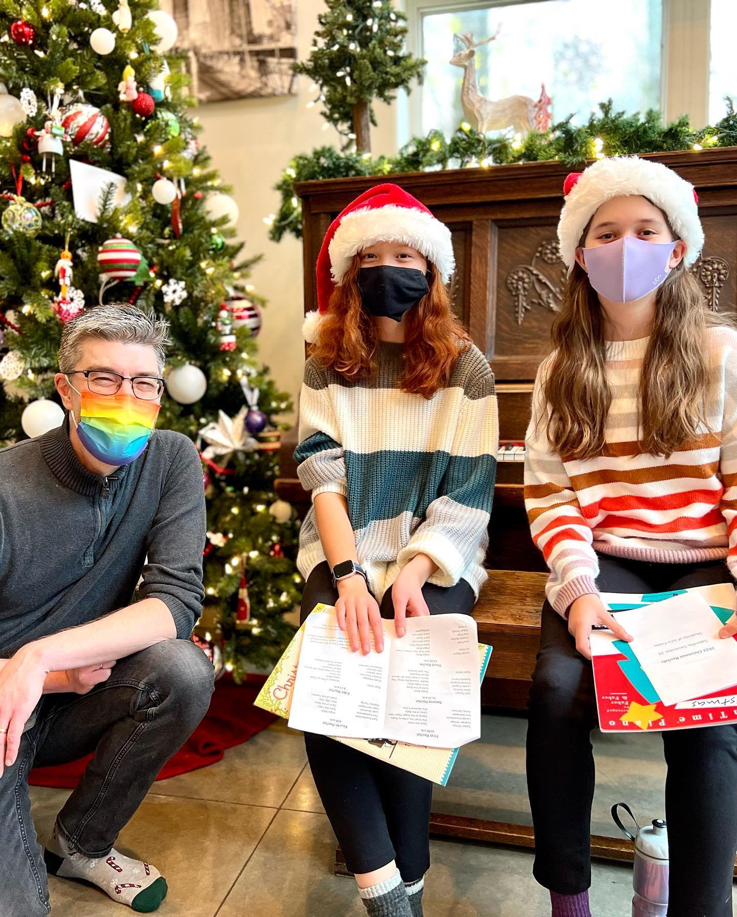 A small group wearing Santa hats and face masks practice singing near a beautifully decorated Christmas tree, holding sheet music. They are in a warm, festive indoor setting.